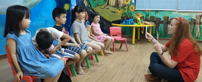 A student is seated on the floor cross-legged, teaching English to Chinese children. 
